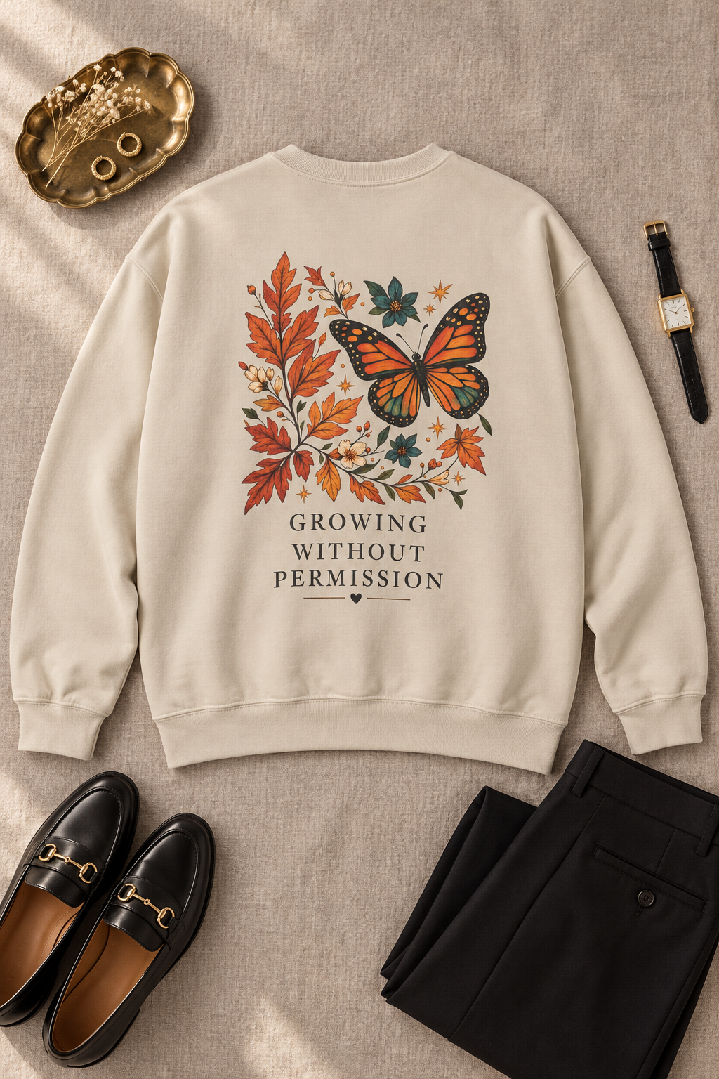 A beige crewneck sweatshirt with a graphic of an orange monarch butterfly surrounded by autumn leaves and the text 'GROWING WITHOUT PERMISSION', styled in a flat lay with black loafers, black trousers, a watch, and a gold tray on a beige fabric background.