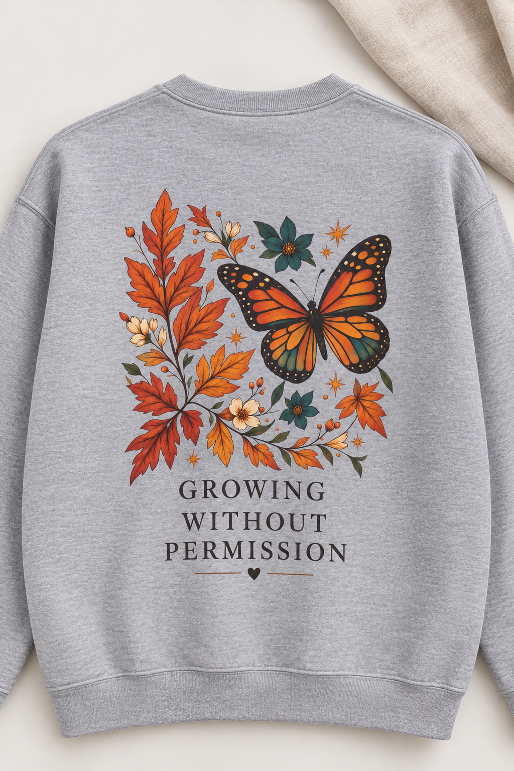 A light heather gray sweatshirt laid flat, featuring a large back graphic with an orange and black butterfly surrounded by autumn leaves and the text 'GROWING WITHOUT PERMISSION' in dark gray.