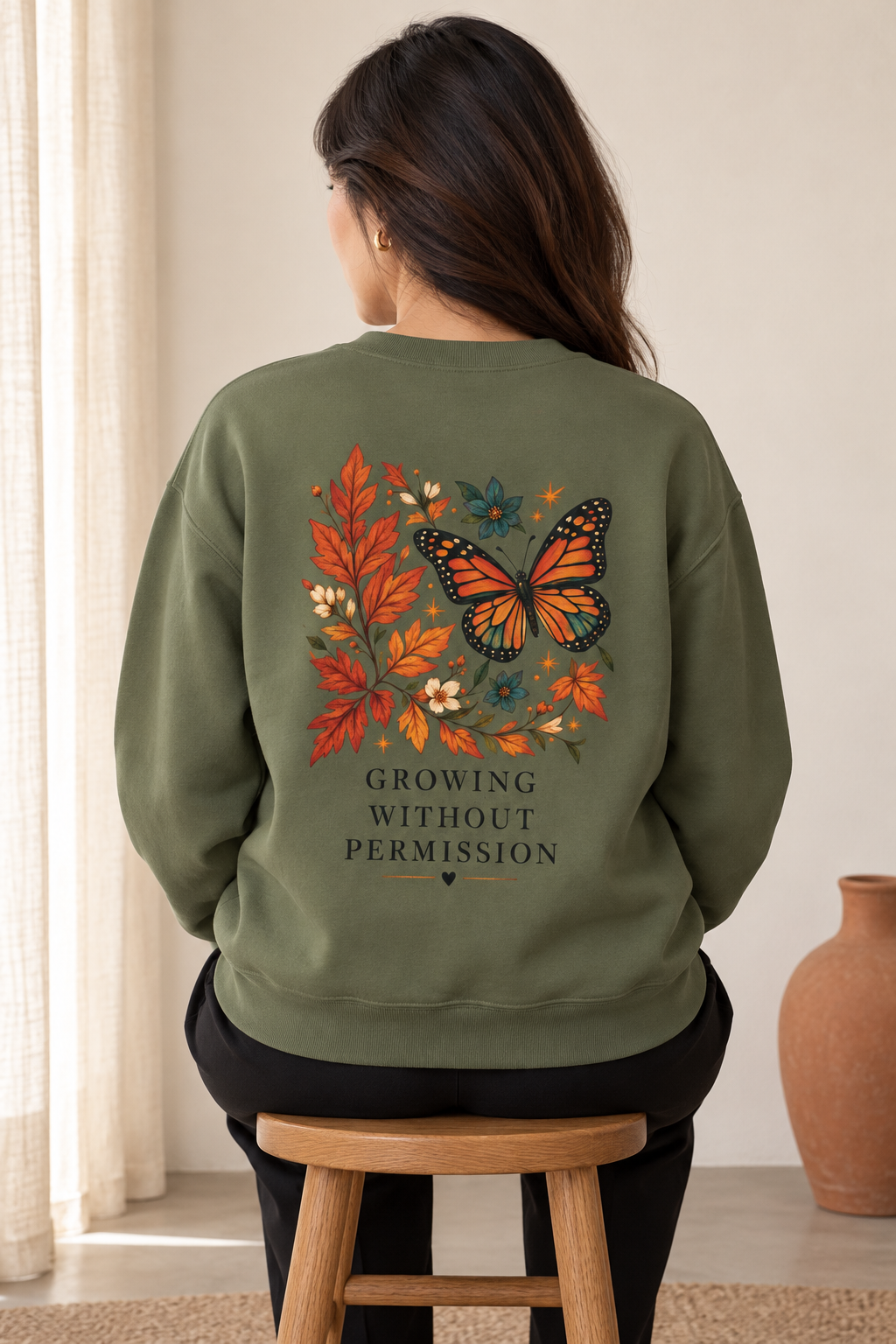 A woman viewed from behind wearing an olive green sweatshirt with a large graphic on the back featuring a monarch butterfly, autumn leaves, flowers, and the text 'GROWING WITHOUT PERMISSION'. She is sitting on a wooden stool in a softly lit indoor setting.