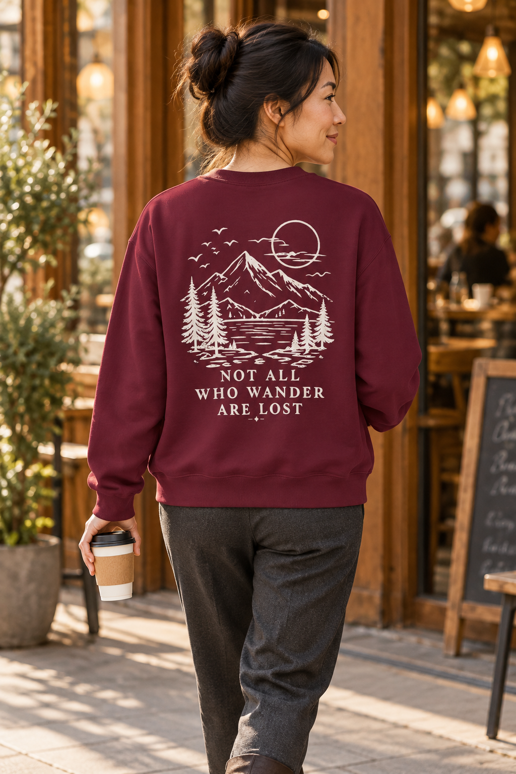 A woman wearing a burgundy crewneck sweatshirt with a cream mountain and lake graphic and the text 'NOT ALL WHO WANDER ARE LOST' on the back, walking in a cafe setting.