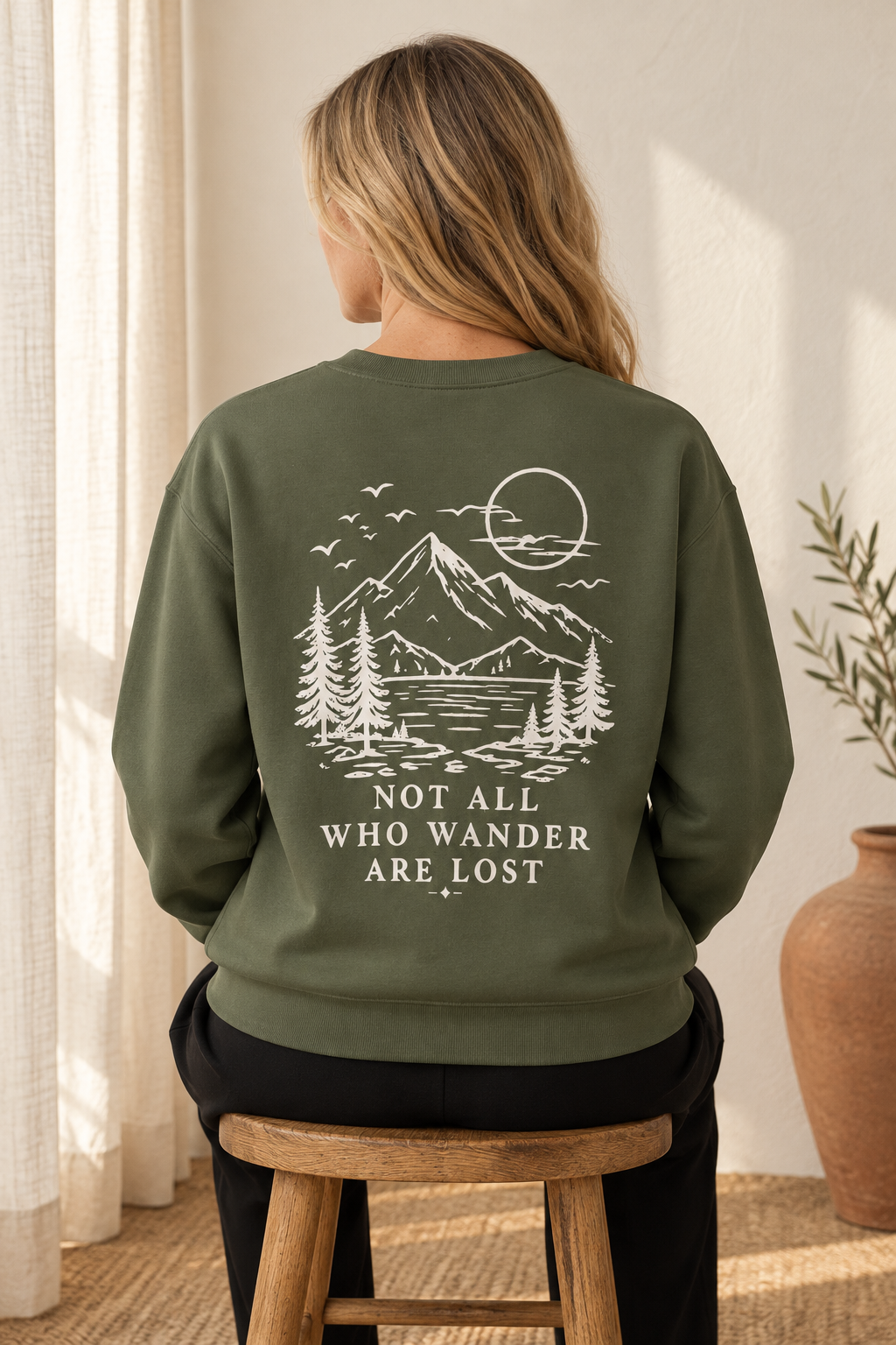 A woman viewed from behind wearing an olive green sweatshirt with a cream graphic of a mountain, lake, trees, and sun, with the text 'NOT ALL WHO WANDER ARE LOST'. She is sitting on a wooden stool in a sunlit room.