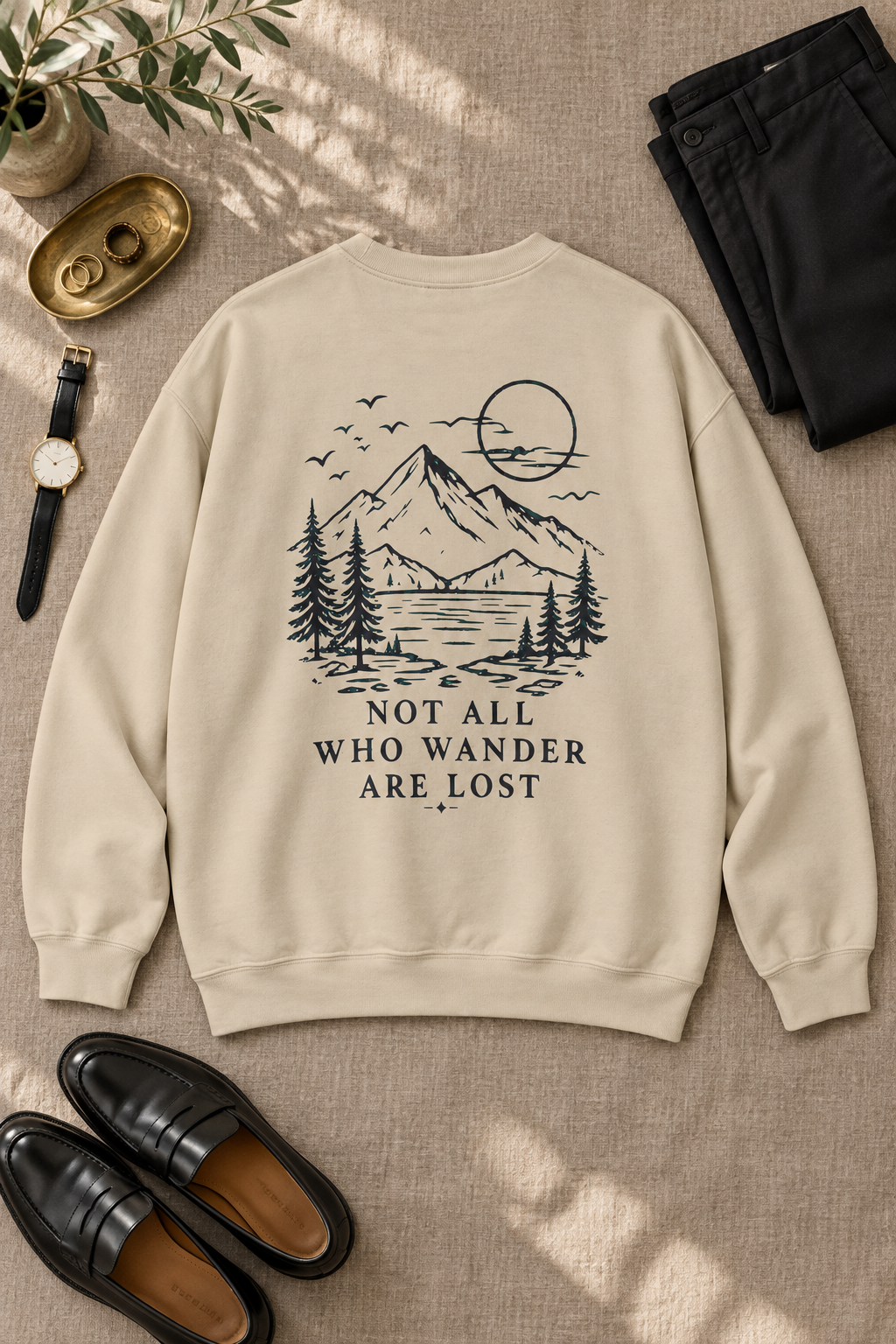 A cream-colored crewneck sweatshirt with a black line art graphic of a mountain, trees, and a sun, with the text 'NOT ALL WHO WANDER ARE LOST' printed on the back. The sweatshirt is styled in a flat-lay arrangement on a textured beige fabric background, surrounded by black pants, black loafers, a watch, and a small plant.