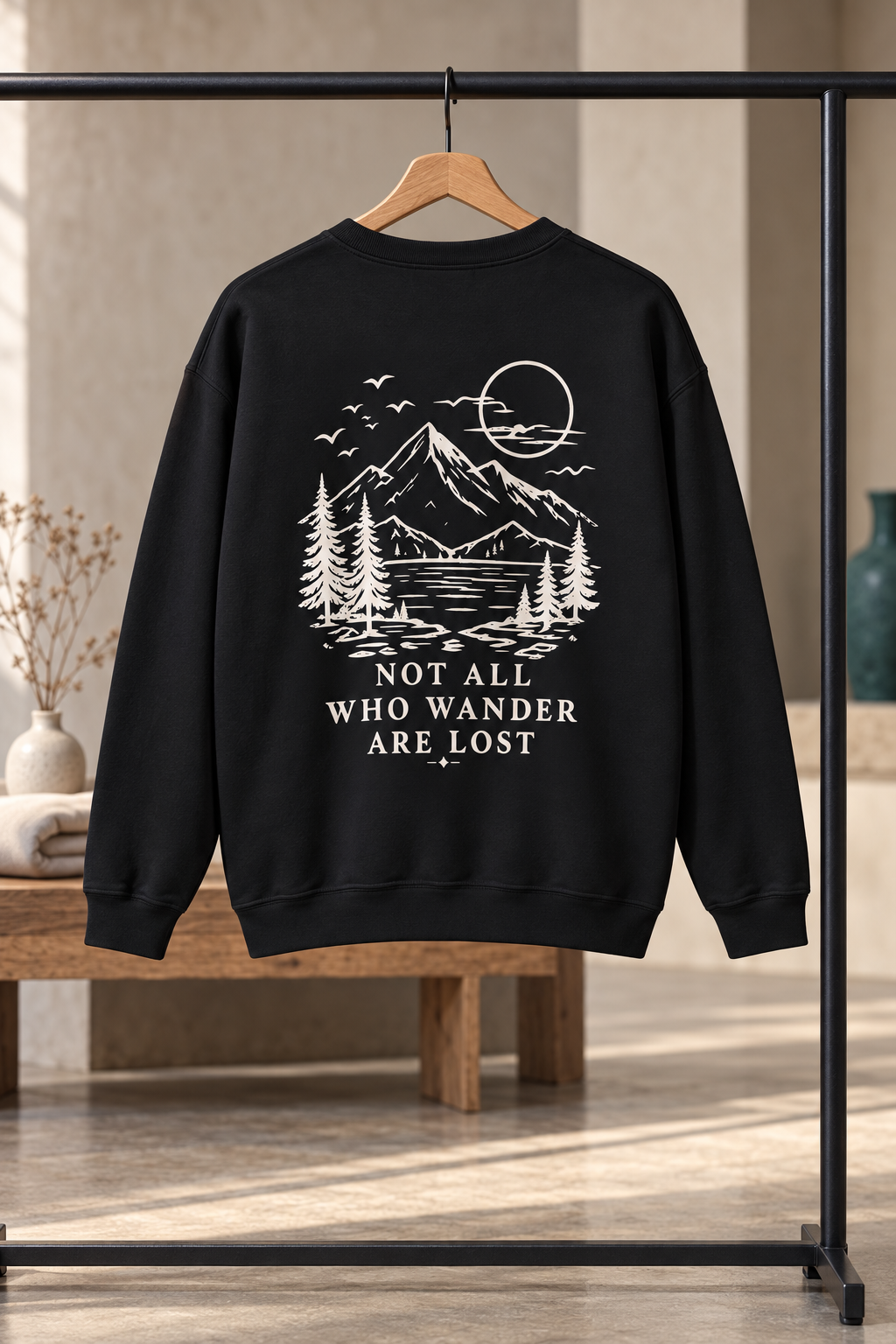 A black crewneck sweatshirt hanging on a wooden hanger from a black clothing rack, featuring a cream graphic of a mountain, pine trees, and a moon with the text 'NOT ALL WHO WANDER ARE LOST'. The background is a softly blurred indoor setting with warm lighting.