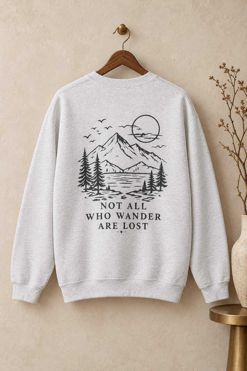 A light heather gray crewneck sweatshirt hanging on a wooden hanger against a textured beige wall. The back of the sweatshirt features a black with a hint of teal line art graphic of a mountain, pine trees, a lake, birds, and a sun, with the text 'NOT ALL WHO WANDER ARE LOST' printed below.