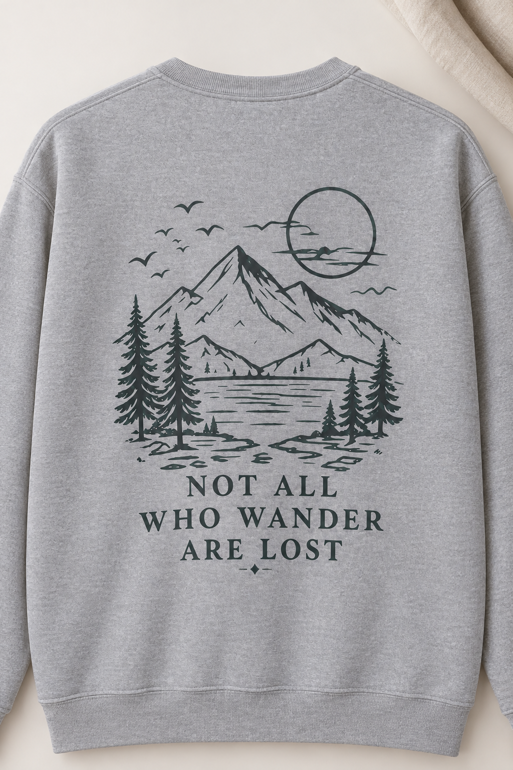 A light heather gray crewneck sweatshirt featuring a black with hint of teal line art graphic of a mountain, lake, pine trees, birds, and a sun, with the text 'NOT ALL WHO WANDER ARE LOST' printed below.