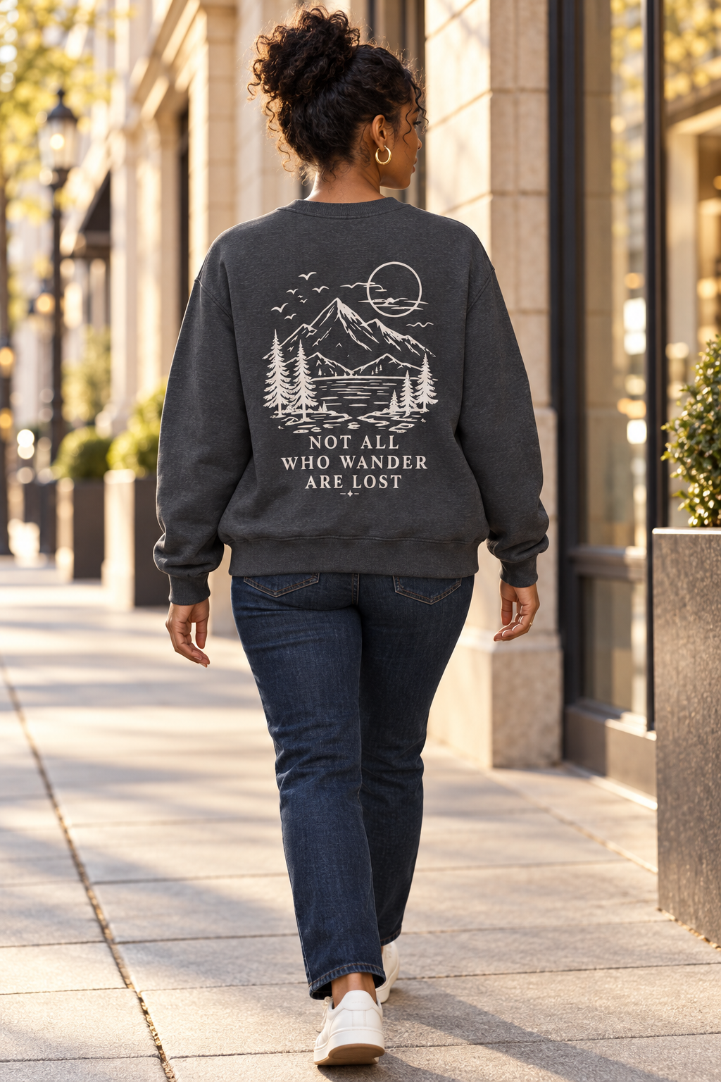 A woman walking on a city sidewalk viewed from behind, wearing a dark charcoal gray crewneck sweatshirt with a cream graphic of a mountain, trees, and the text 'NOT ALL WHO WANDER ARE LOST', paired with dark blue jeans and white sneakers.