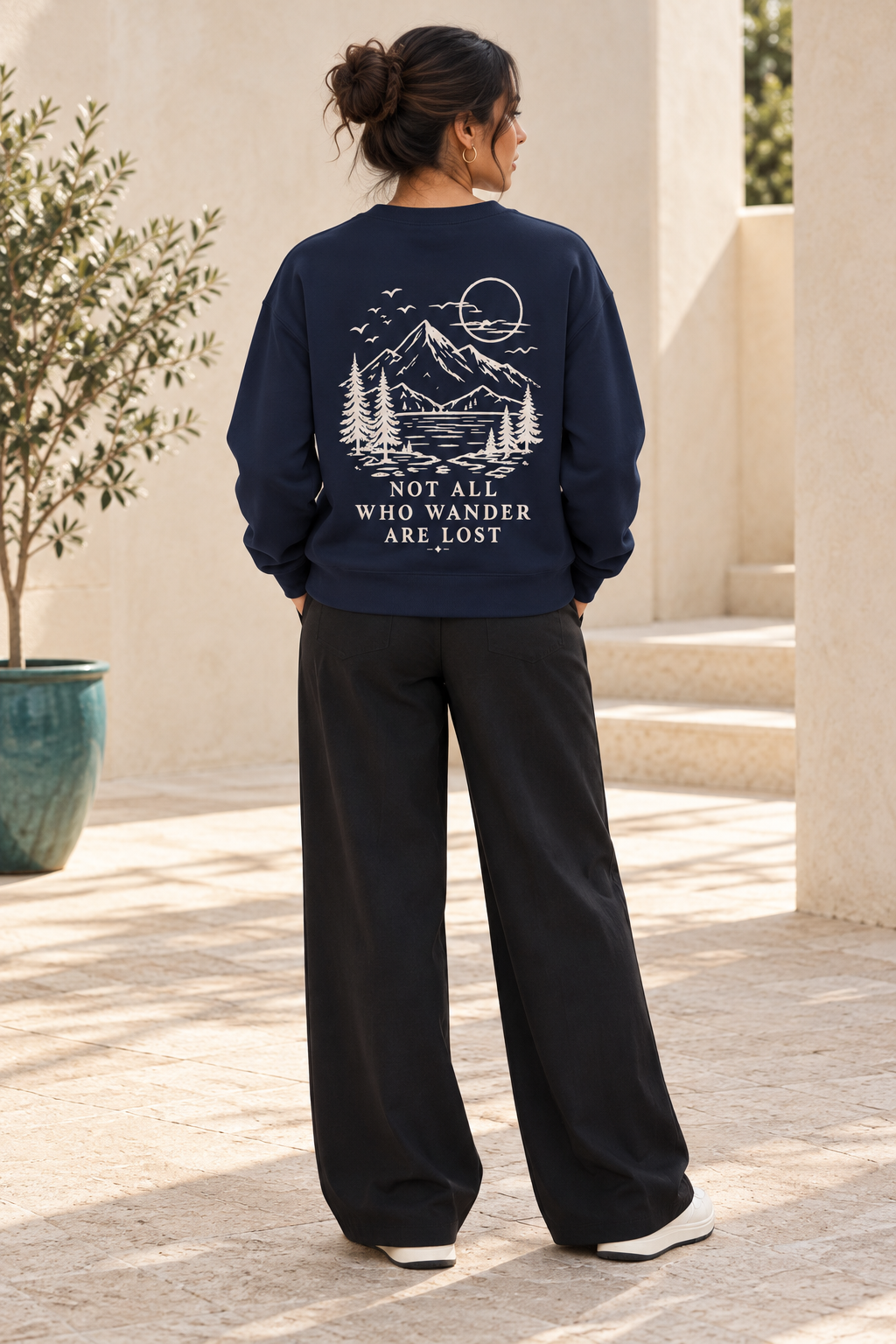 A woman viewed from behind wearing a navy blue sweatshirt with a cream mountain and lake graphic and the text 'NOT ALL WHO WANDER ARE LOST' on the back, paired with black wide-leg pants in an outdoor patio setting.