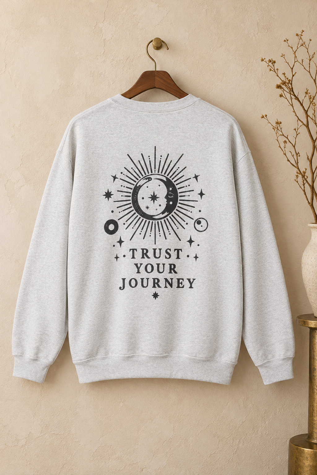 A light heather gray crewneck sweatshirt hanging on a wooden hanger against a textured beige wall. The back of the sweatshirt features a black graphic of a sun and moon with stars and the text 'TRUST YOUR JOURNEY'.