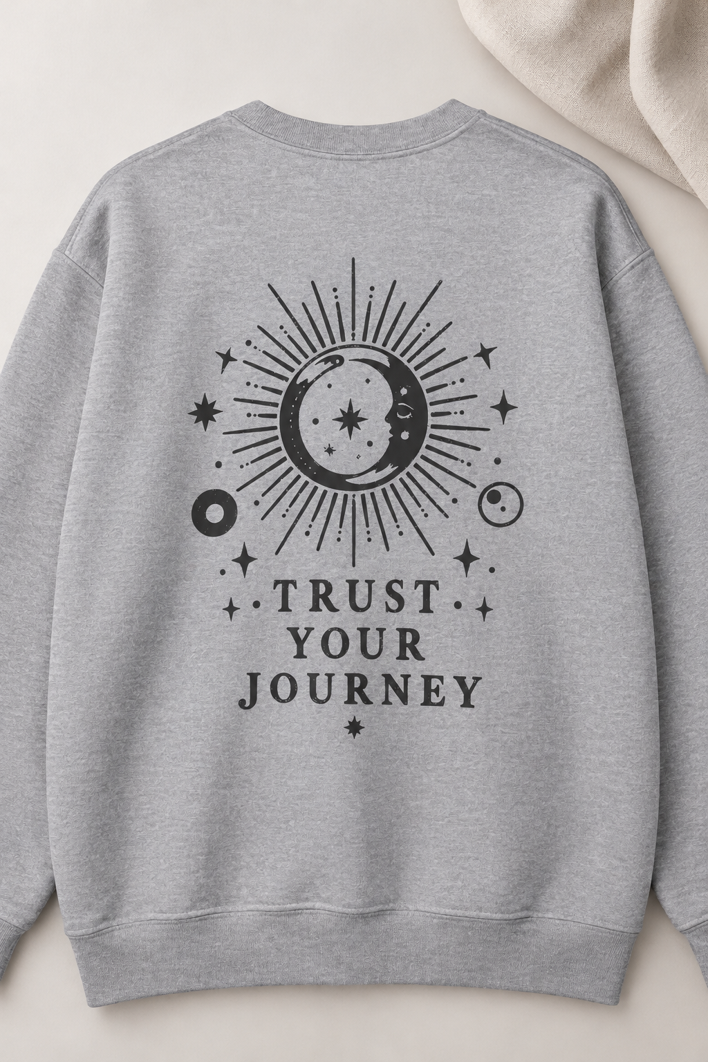 A light heather gray crewneck sweatshirt featuring a black celestial graphic on the back with a sun, moon, stars, and the text 'TRUST YOUR JOURNEY'.