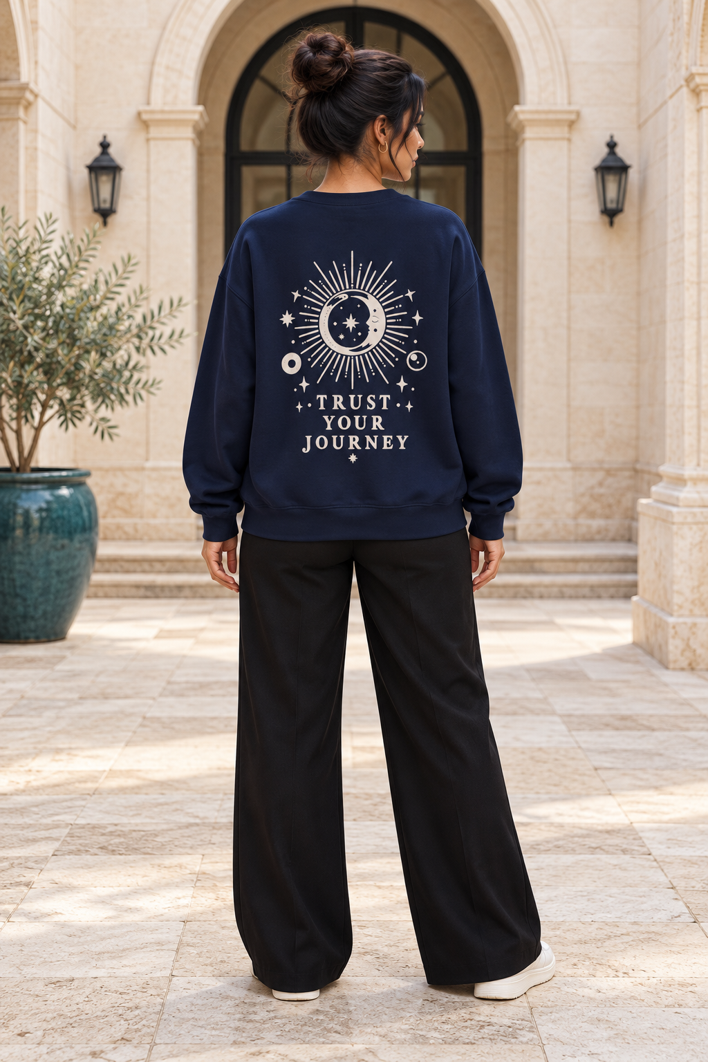 A woman viewed from behind wearing a navy blue sweatshirt with a cream celestial graphic and the text 'TRUST YOUR JOURNEY' on the back, paired with black wide-leg pants, standing in front of a stone building.