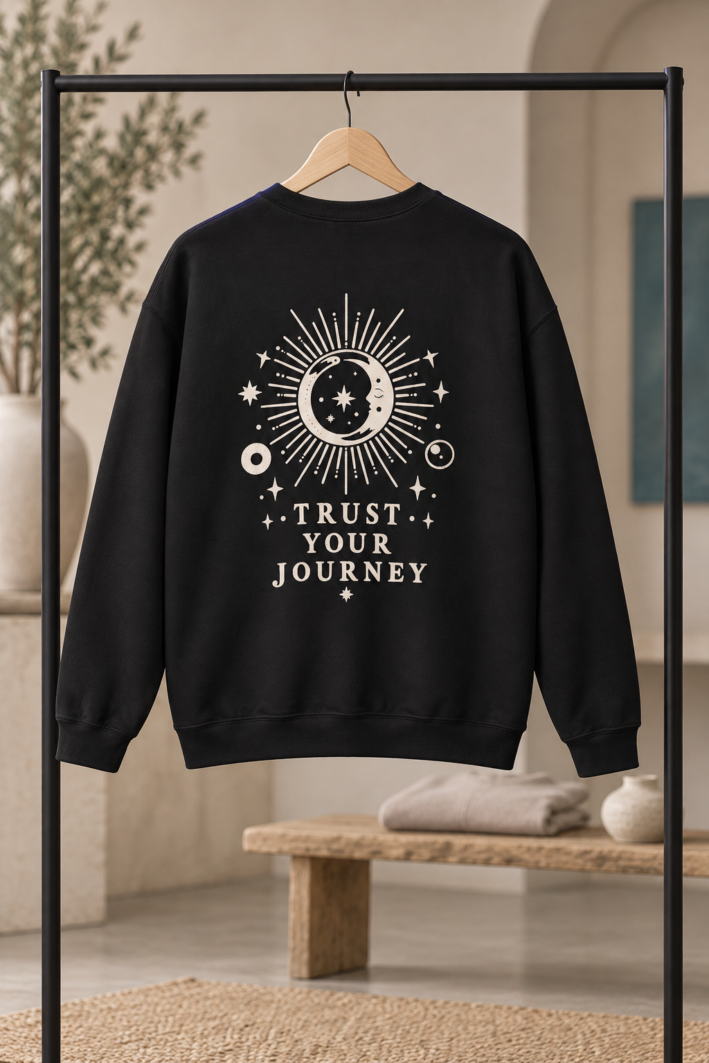 A black crewneck sweatshirt hanging on a wooden hanger from a black clothing rack, featuring a cream celestial graphic of a sun, moon, and stars with the text 'TRUST YOUR JOURNEY' on the back. The background is a softly blurred warm-toned interior with a bench and plants.