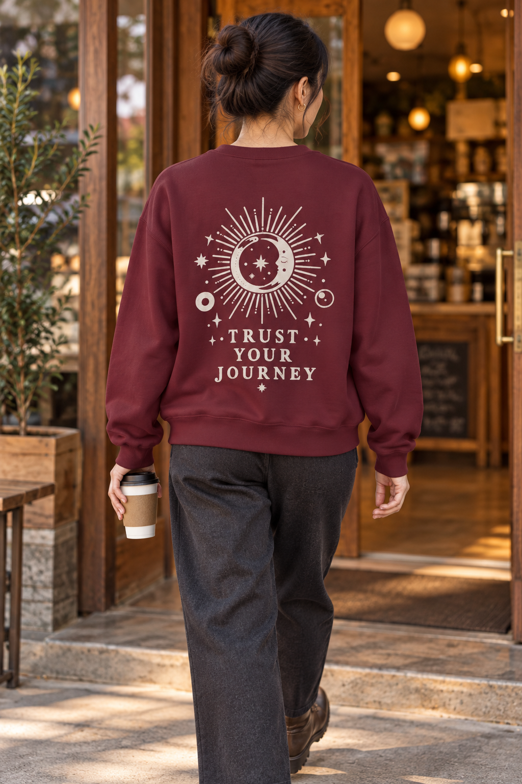 A woman viewed from behind walking through a cafe doorway, wearing a burgundy crewneck sweatshirt with a cream celestial graphic and the words 'TRUST YOUR JOURNEY' on the back, paired with dark gray sweatpants and holding a coffee cup.