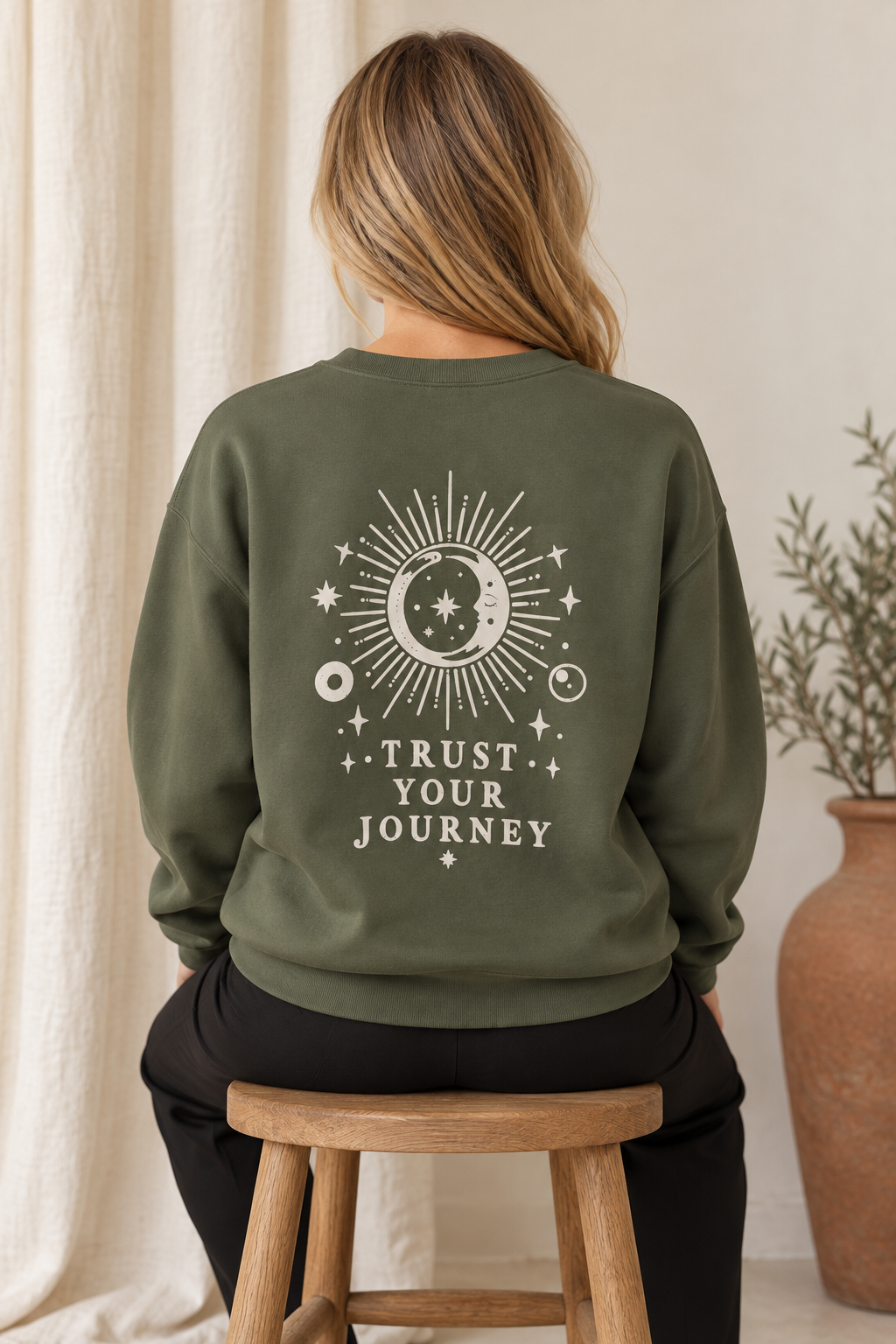A woman viewed from behind wearing an olive green sweatshirt with a cream celestial sun and moon graphic and the text 'TRUST YOUR JOURNEY' on the back, seated on a wooden stool in a softly lit indoor setting.