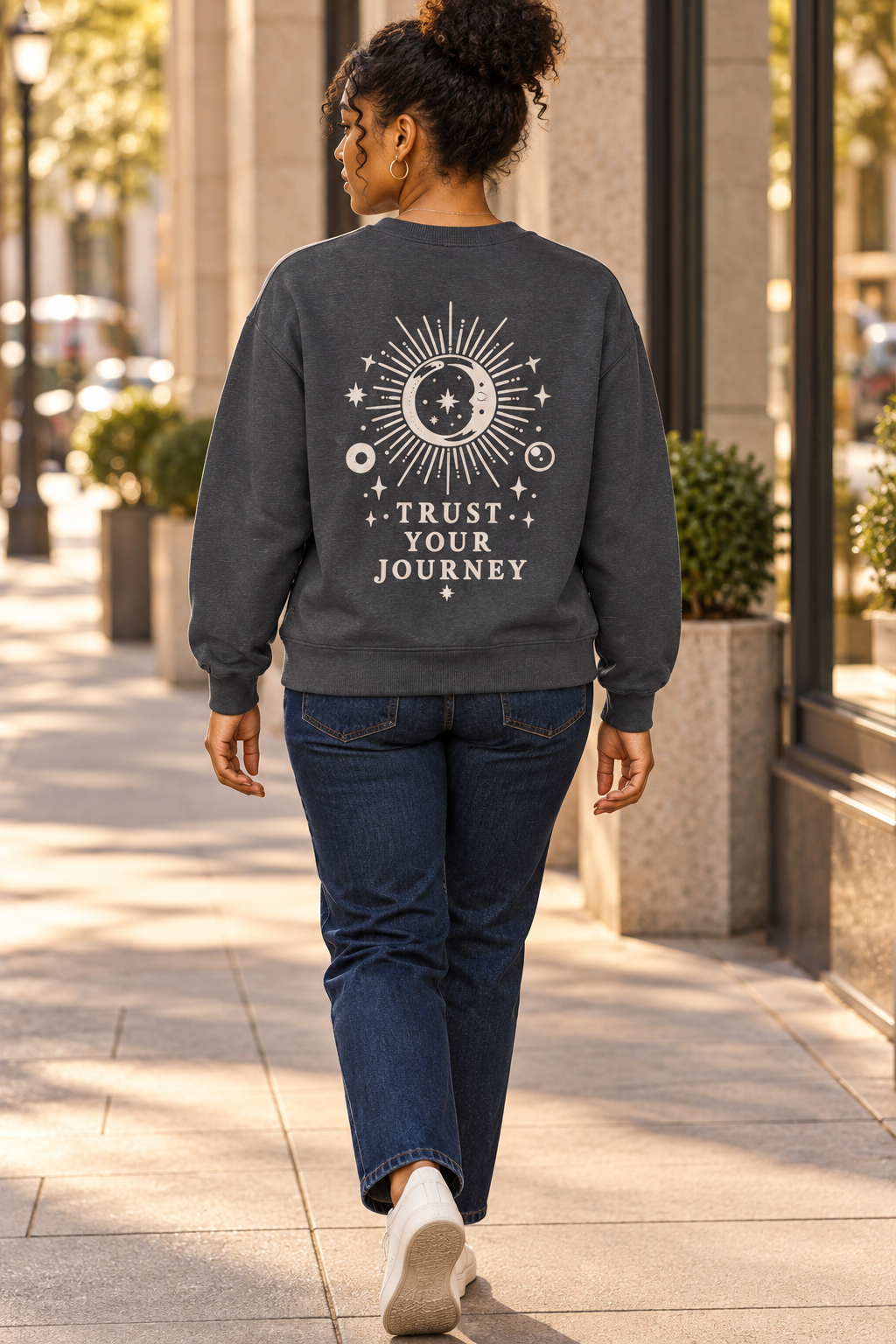 A woman viewed from behind walking on a city sidewalk, wearing a dark charcoal gray sweatshirt with a cream celestial graphic and the words 'TRUST YOUR JOURNEY' on the back, paired with dark blue jeans and white sneakers.