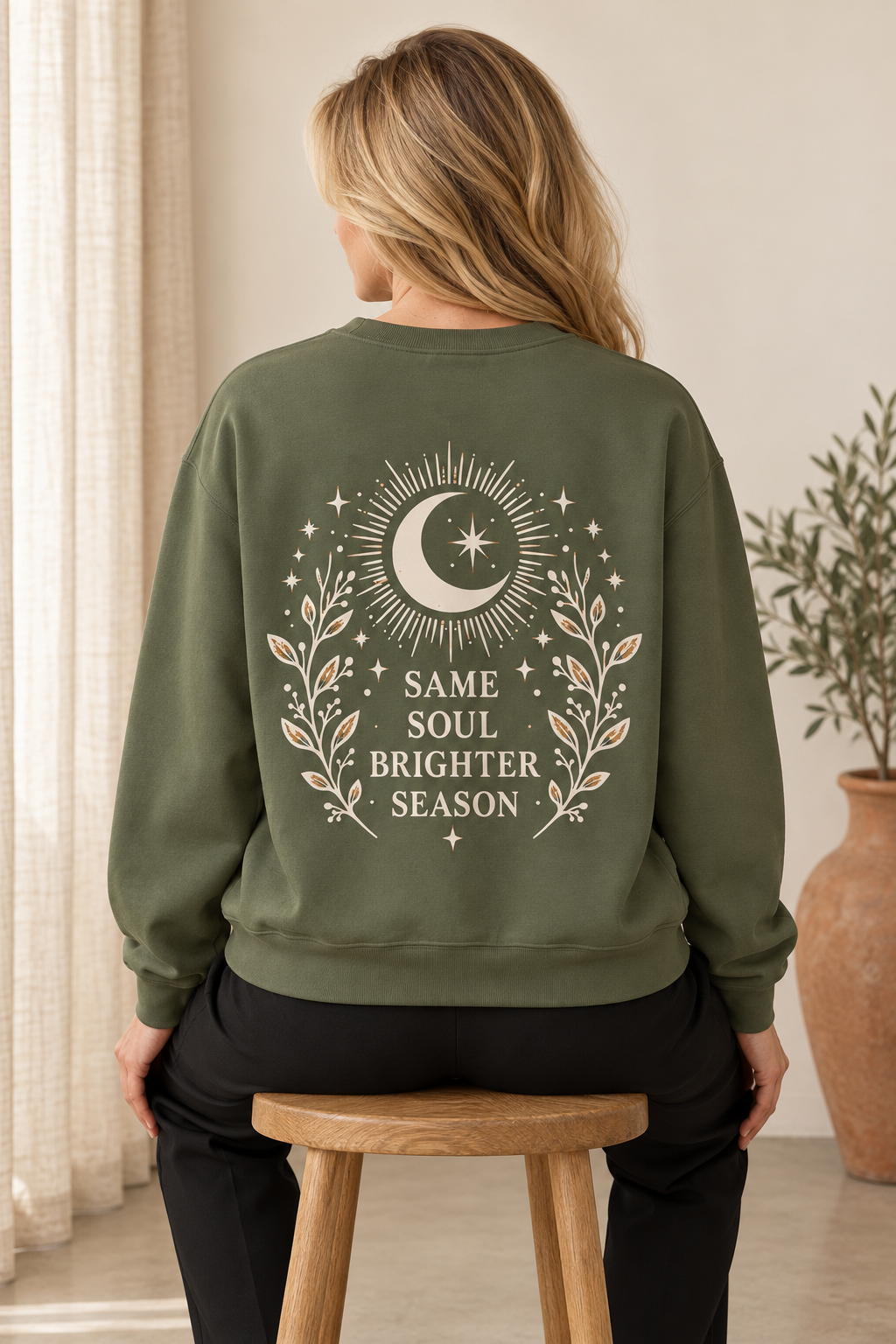 A woman viewed from behind wearing an olive green sweatshirt with a cream-colored celestial graphic on the back. The graphic features a crescent moon, sunburst, stars, and the text 'SAME SOUL BRIGHTER SEASON.