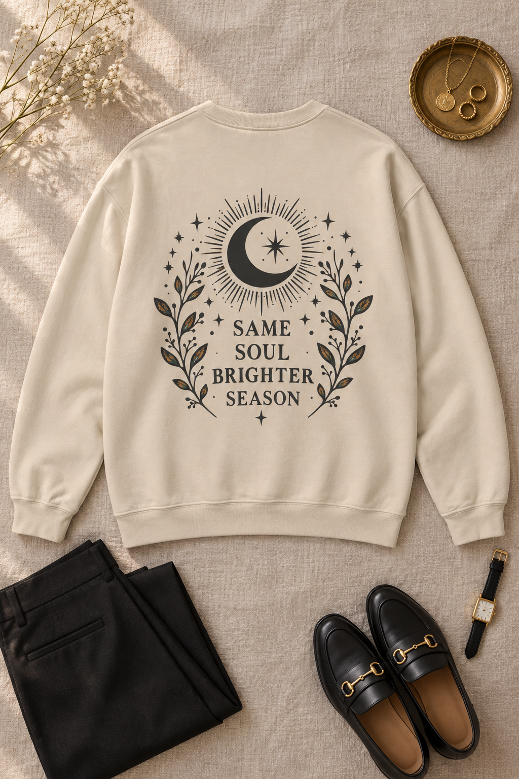 A beige crewneck sweatshirt with a black bohemian-style graphic on the back featuring a crescent moon, stars, and the text 'SAME SOUL BRIGHTER SEASON'. The sweatshirt is styled in a flat-lay arrangement on a textured beige fabric background, accompanied by black trousers, black leather loafers with gold hardware, a watch, and dried flowers.