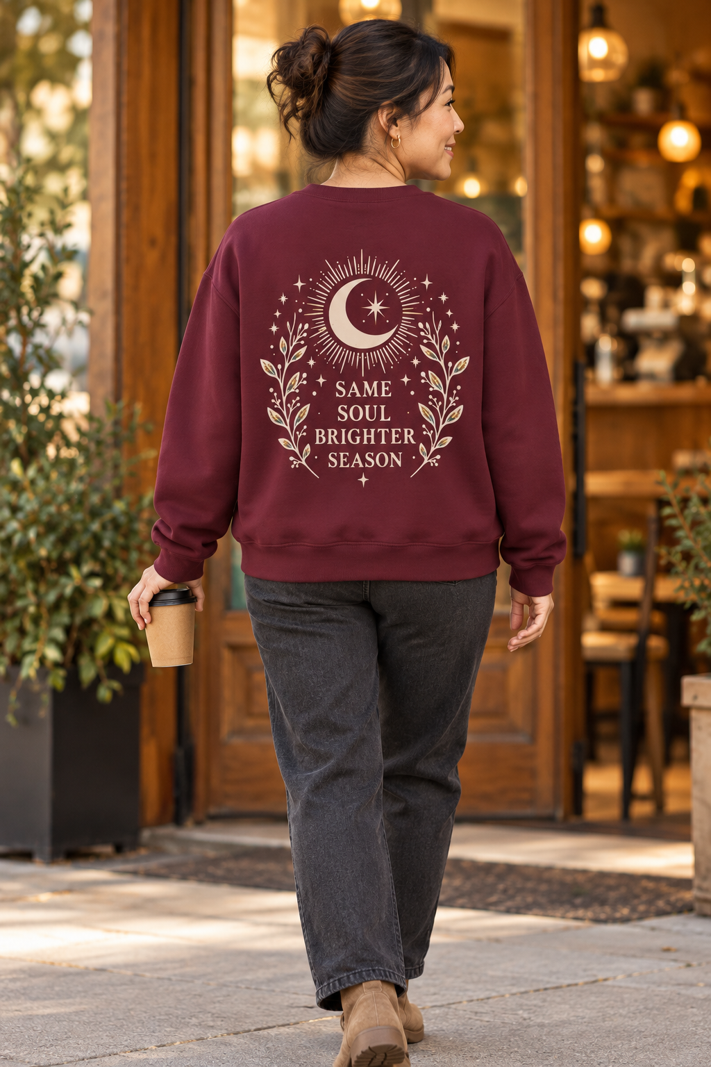 A woman viewed from behind walking outdoors in front of a warm storefront, wearing a burgundy crewneck sweatshirt with a large cream moon and floral graphic and the text 'SAME SOUL BRIGHTER SEASON' on the back.