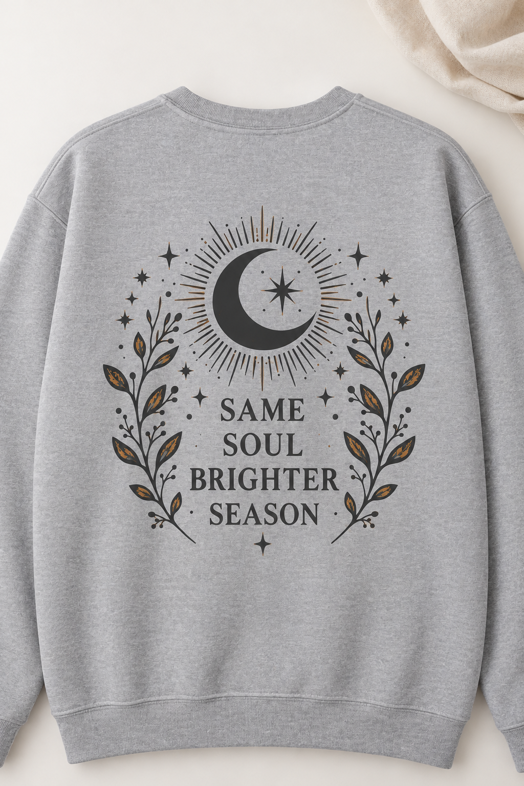 A light heather gray crewneck sweatshirt shown from the back, featuring a dark graphic print of a crescent moon, stars, and leafy branches with the text 'SAME SOUL BRIGHTER SEASON'.