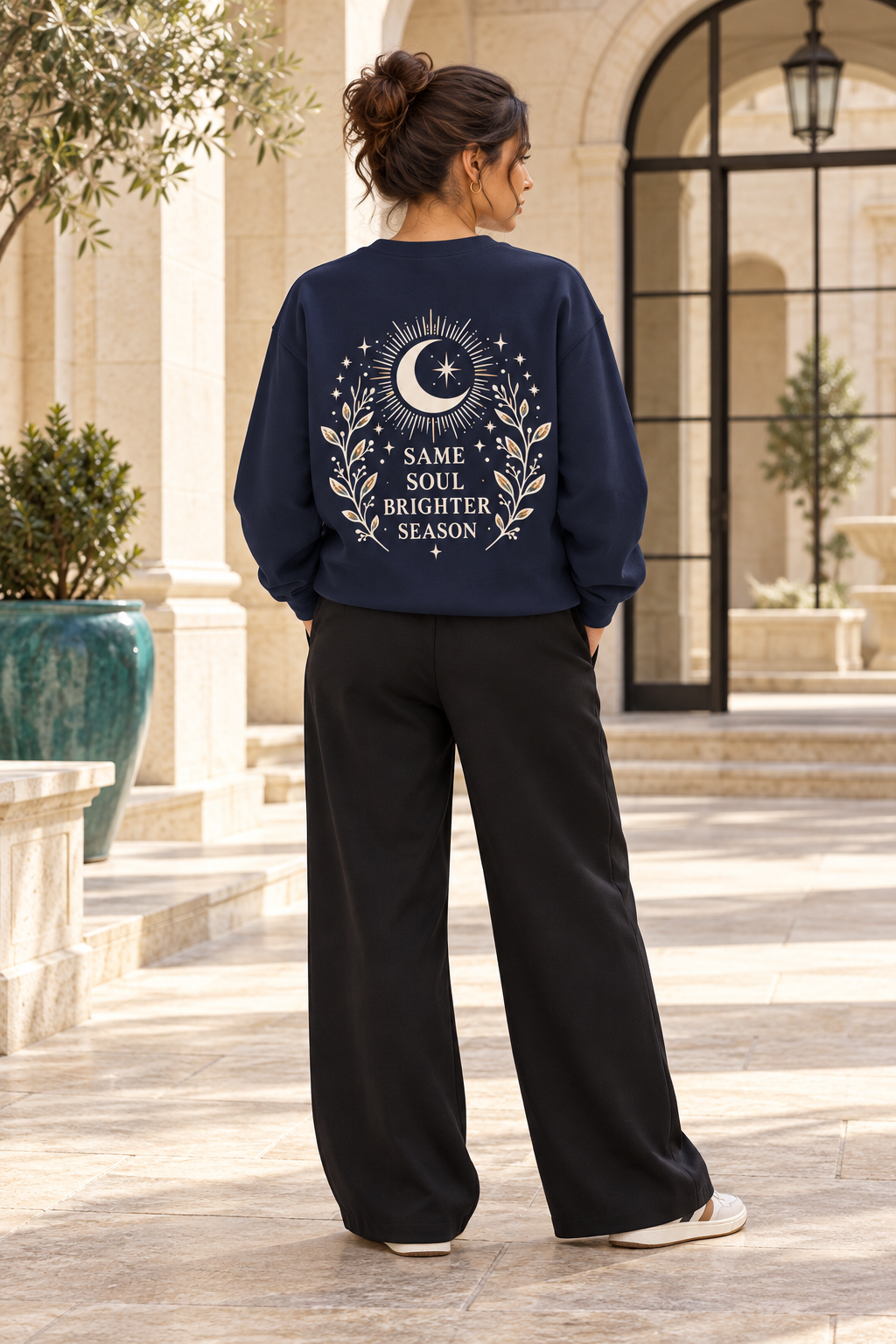 A woman viewed from behind wearing a navy blue sweatshirt with a cream celestial graphic and the text 'SAME SOUL BRIGHTER SEASON' on the back, paired with black wide-leg pants in an outdoor courtyard setting.