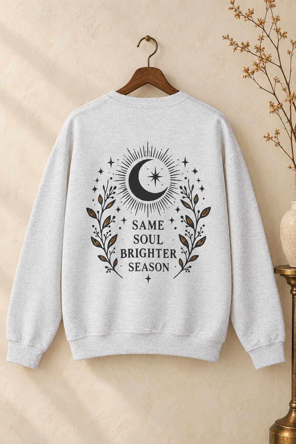 A light heather gray crewneck sweatshirt hanging on a wooden hanger against a warm beige wall. The back of the sweatshirt features a charcoal graphic print of a crescent moon surrounded by stars and leafy branches, with the text 'SAME SOUL BRIGHTER SEASON'.