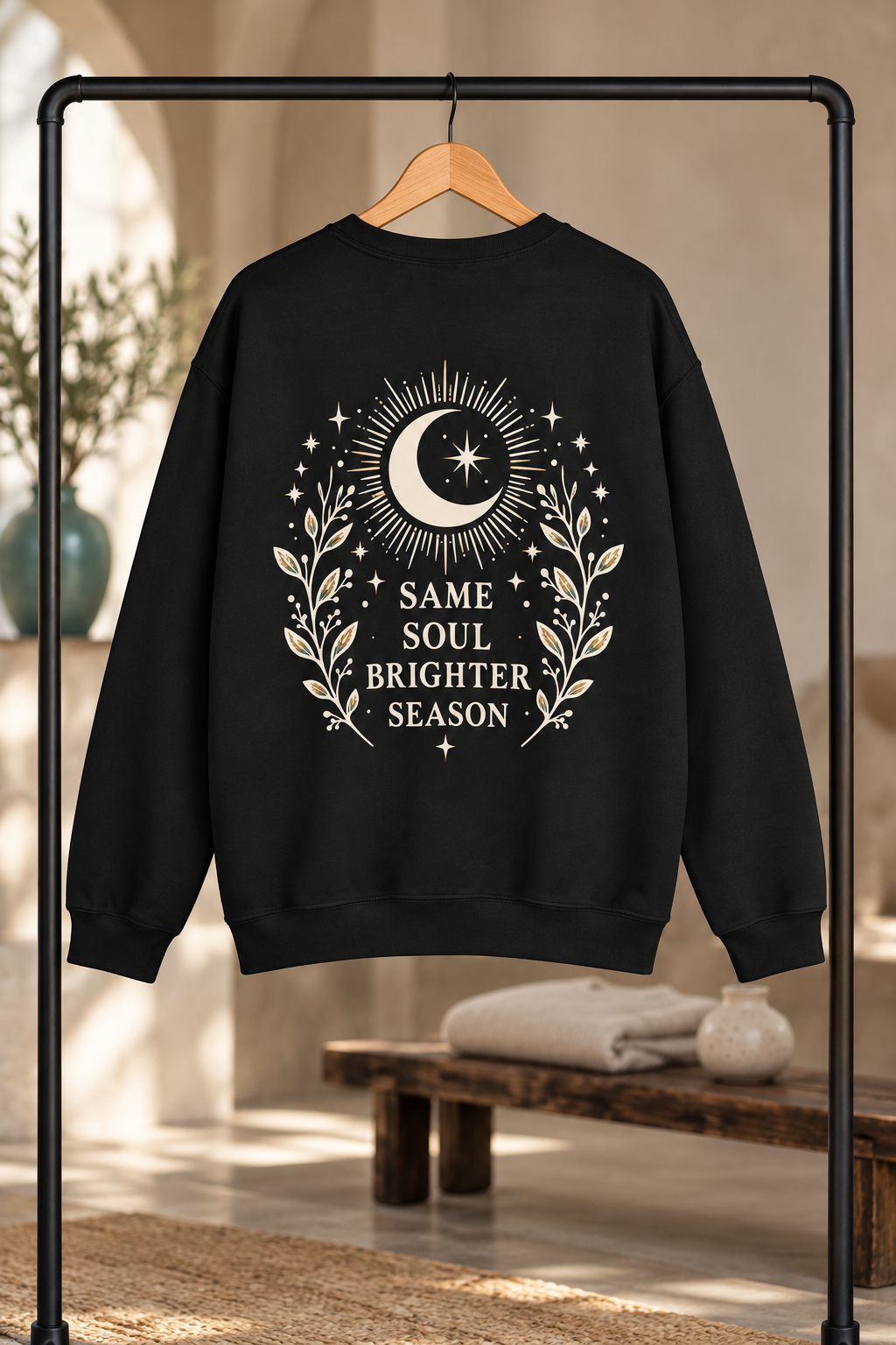 A black crewneck sweatshirt hanging on a wooden hanger from a black clothing rack, featuring a cream graphic of a crescent moon, stars, and a leafy wreath with the text 'SAME SOUL BRIGHTER SEASON'. The background is a softly blurred warm-toned interior with a bench and folded blanket.