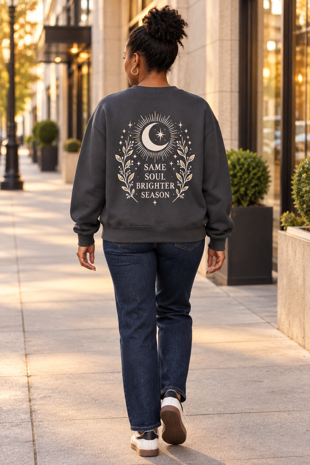 A woman viewed from behind walking on a city sidewalk, wearing a dark charcoal gray sweatshirt with a large cream graphic on the back featuring a crescent moon, stars, and the text 'SAME SOUL BRIGHTER SEASON'. She is also wearing dark blue jeans and white sneakers.