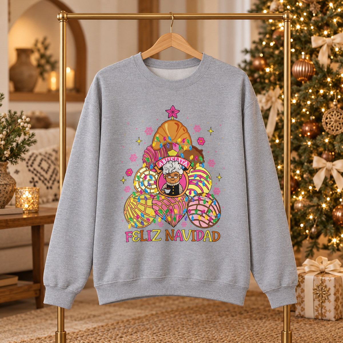 A light heather gray crewneck sweatshirt hanging on a wooden hanger on a gold clothing rack, set against a cozy Christmas tree and holiday living room background. The sweatshirt features a colorful colorful concha christmas tree with abuelita in the middle and the text 'FELIZ NAVIDAD' under the tree.