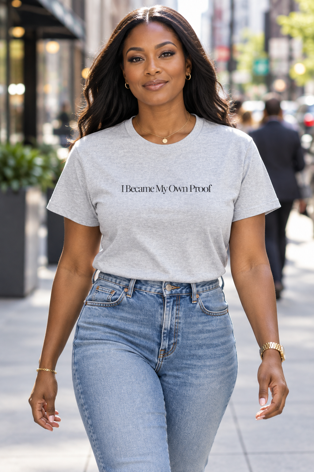 A woman walking on a city street wearing a light gray crewneck t-shirt with the text 'I Became My Own Proof' printed in black across the chest, paired with light-wash blue jeans.