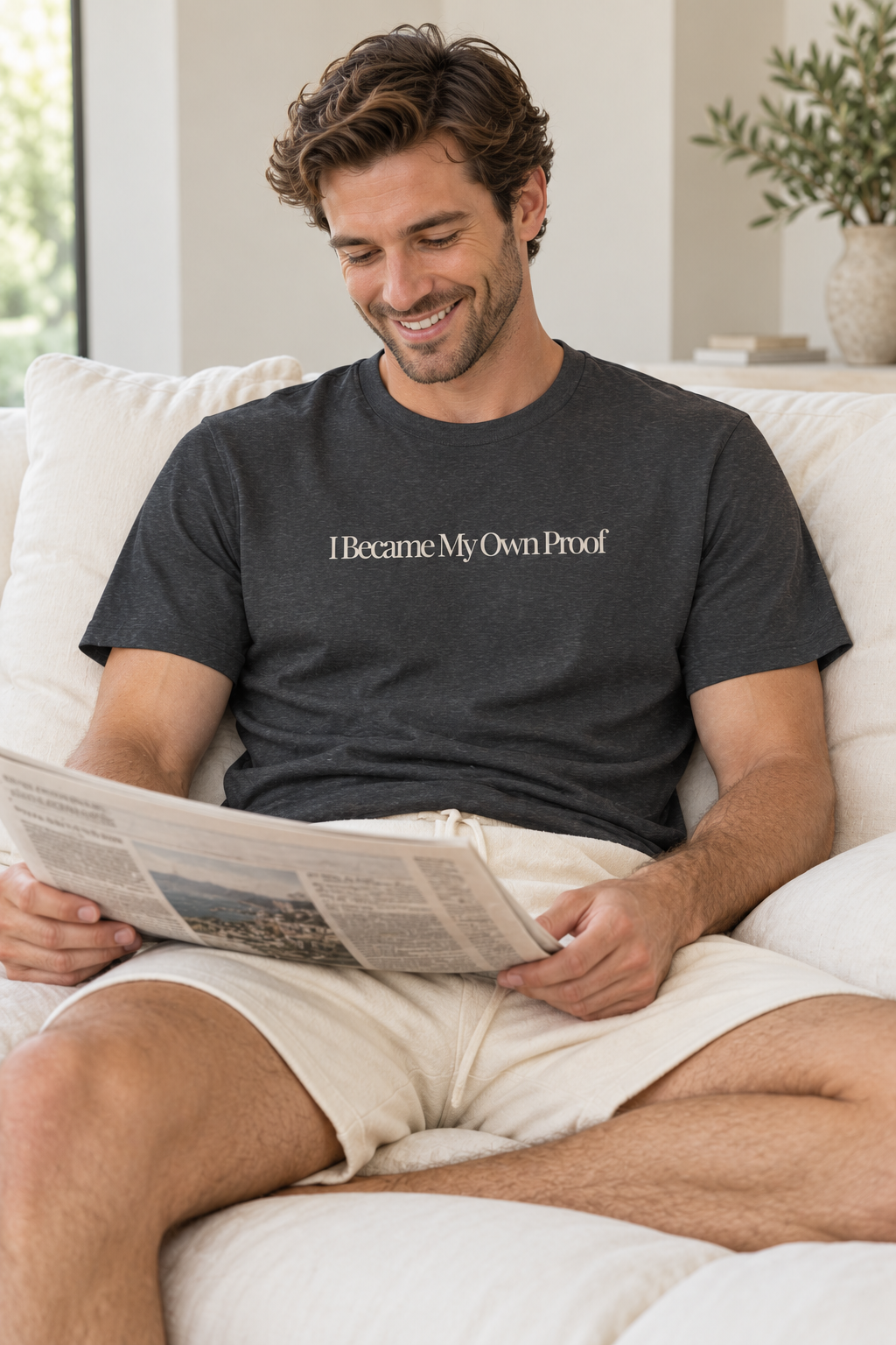 A man sitting on a white couch reading a newspaper, wearing a dark charcoal gray t-shirt with the text 'I Became My Own Proof' in cream, paired with beige shorts.