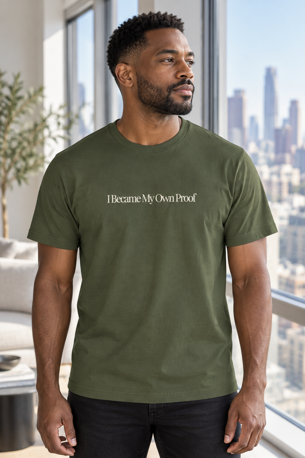 A man wearing an military green crewneck t-shirt with the text 'I Became My Own Proof' printed in cream across the chest, standing in a modern room with a city skyline visible through the window behind him.