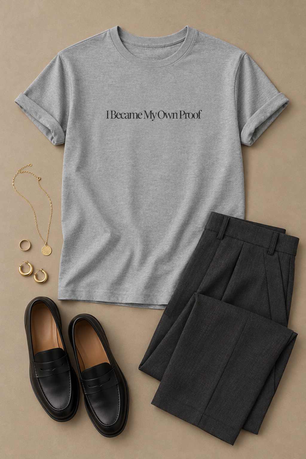 A styled flat-lay featuring a sport gray short-sleeve t-shirt with the text 'I Became My Own Proof' printed in charcoal across the chest. The shirt is arranged alongside dark charcoal gray tailored trousers, black leather loafers, a gold chain necklace, and gold hoop earrings on a solid beige background.