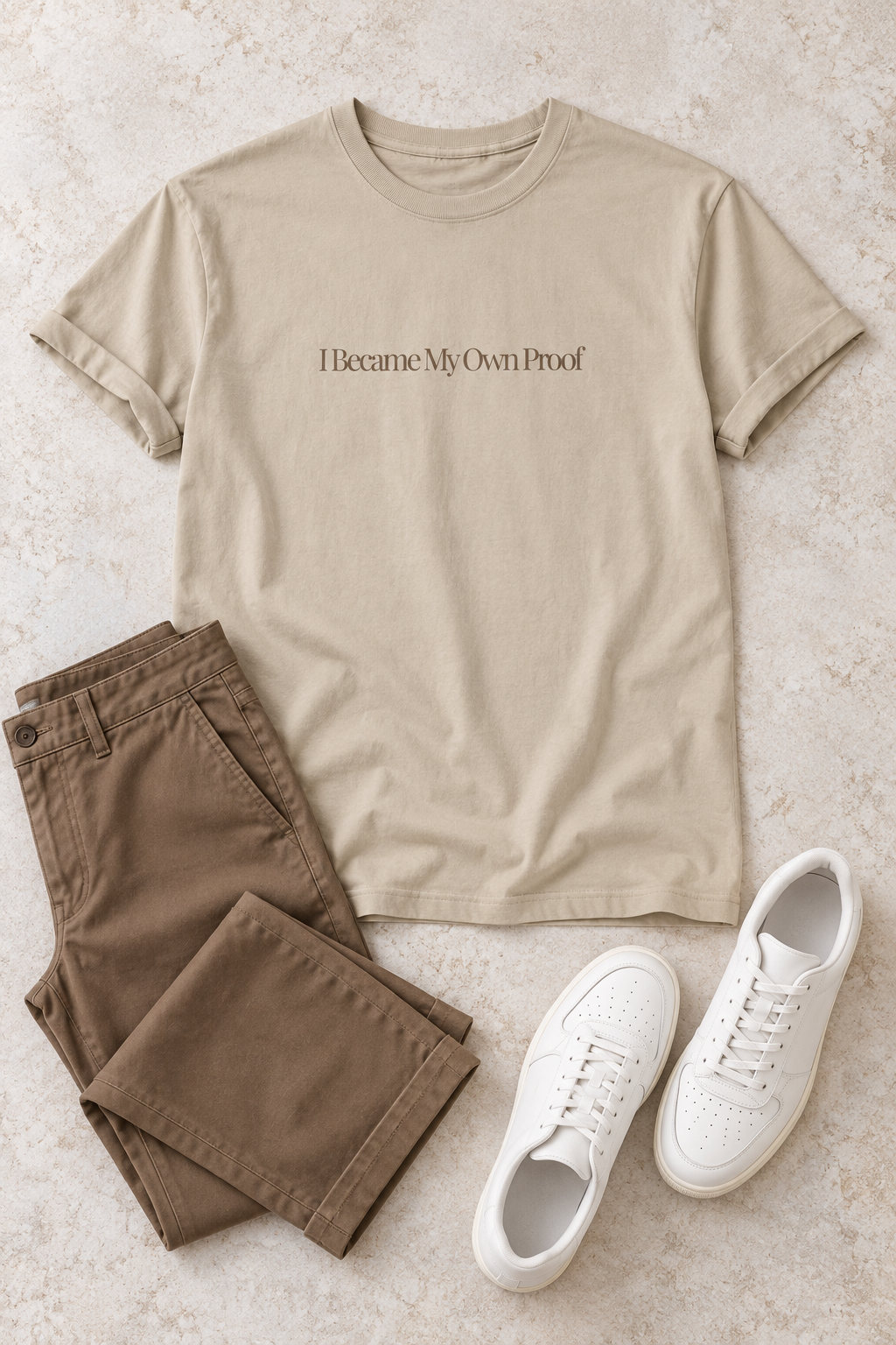 A beige graphic t-shirt with the text 'I Became My Own Proof' printed in expresso across the chest, styled in a flat lay with brown pants and white sneakers on a textured beige background.