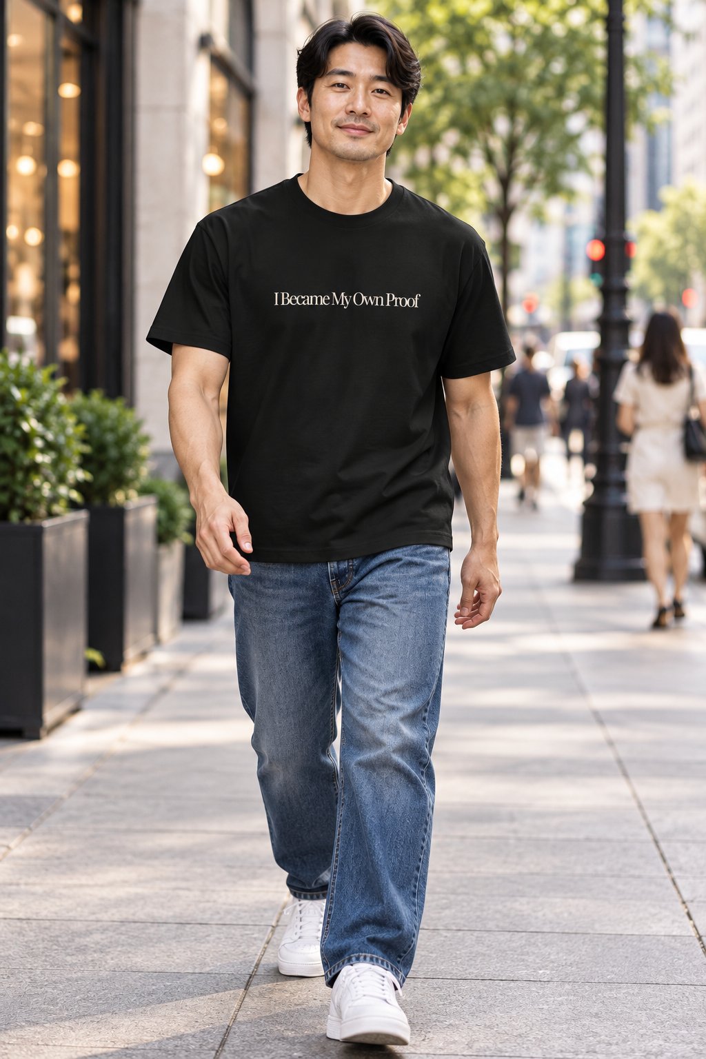 A man walking on a city sidewalk wearing a black crewneck t-shirt with the text 'I Became My Own Proof' printed in cream across the chest, paired with blue jeans and white sneakers.